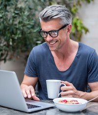 Mann sitzt mit Kaffe und Müsli am Laptop